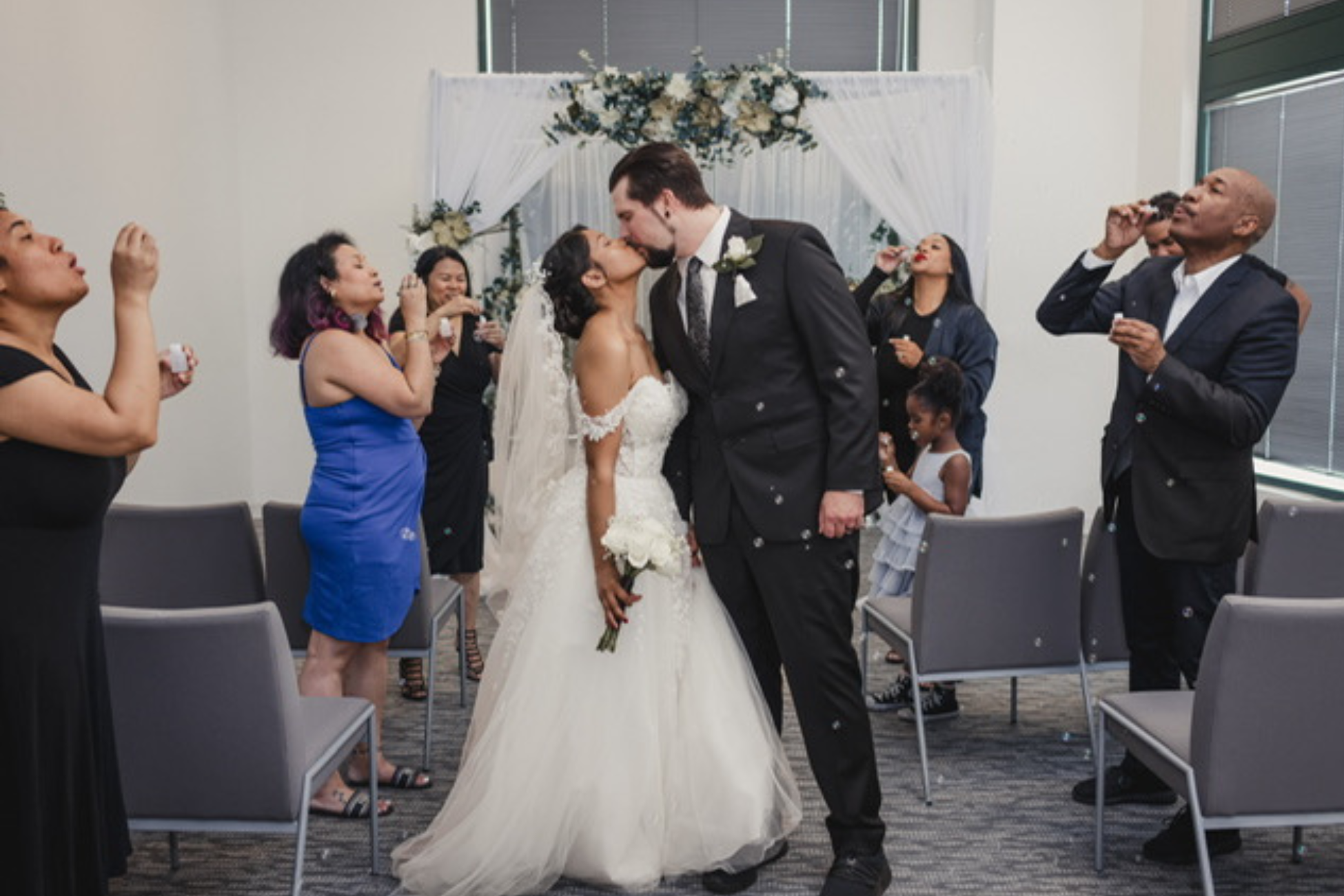 A bride and groom kiss in a wedding ceremony room adorned with a floral arch. Guests on either side blow bubbles, adding a joyful and celebratory atmosphere.