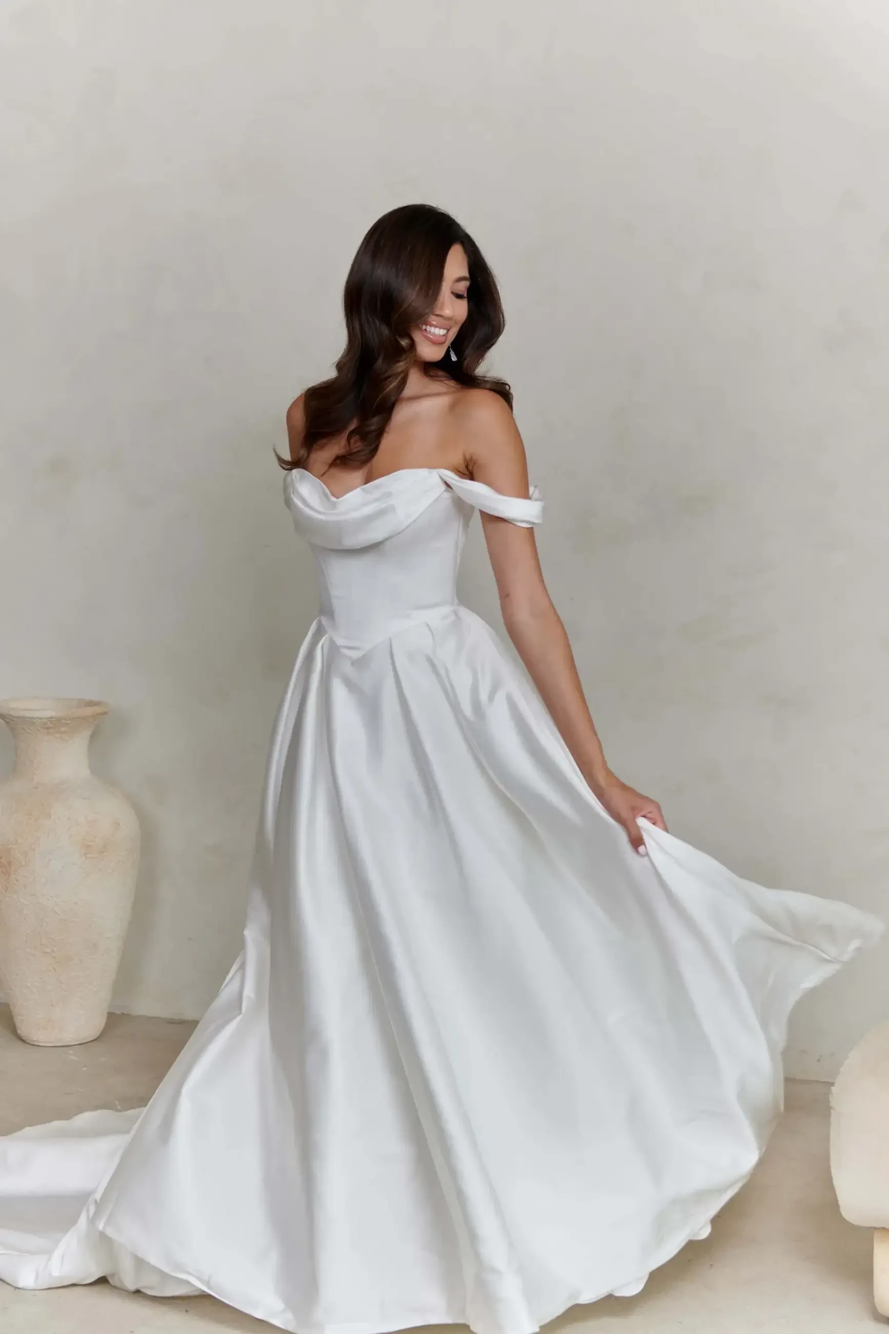 A woman in an elegant white off-shoulder wedding dress smiles as she holds the skirt, standing in a softly lit room with a plain wall and vases.
