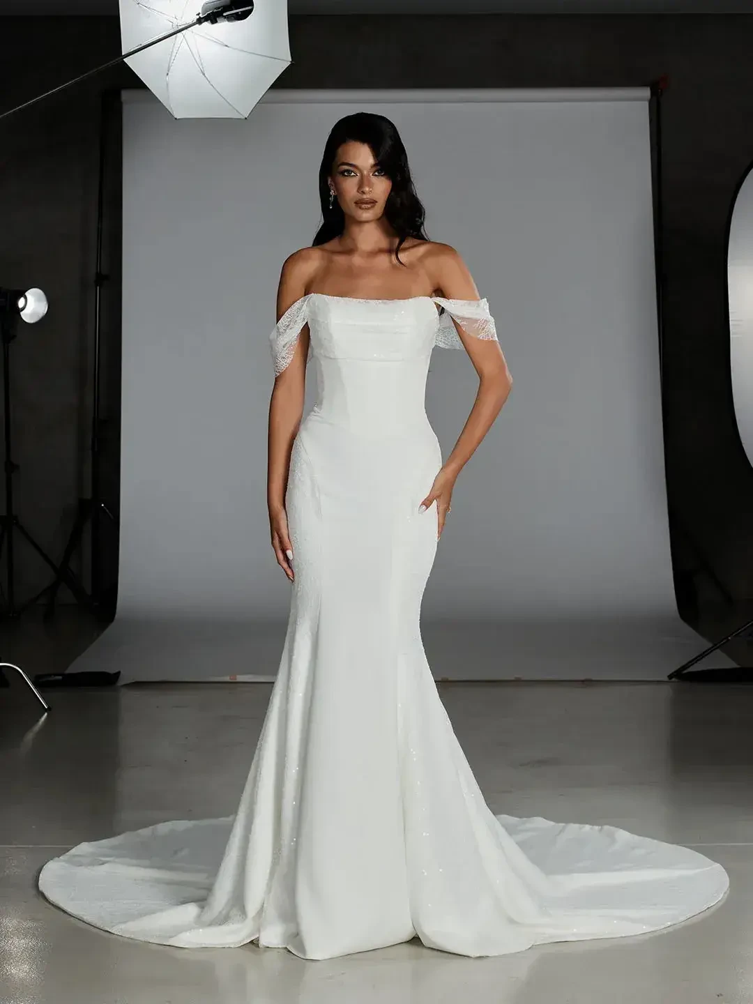 A woman in an elegant, off-the-shoulder white gown stands in a photography studio. She appears confident, surrounded by professional lighting equipment.