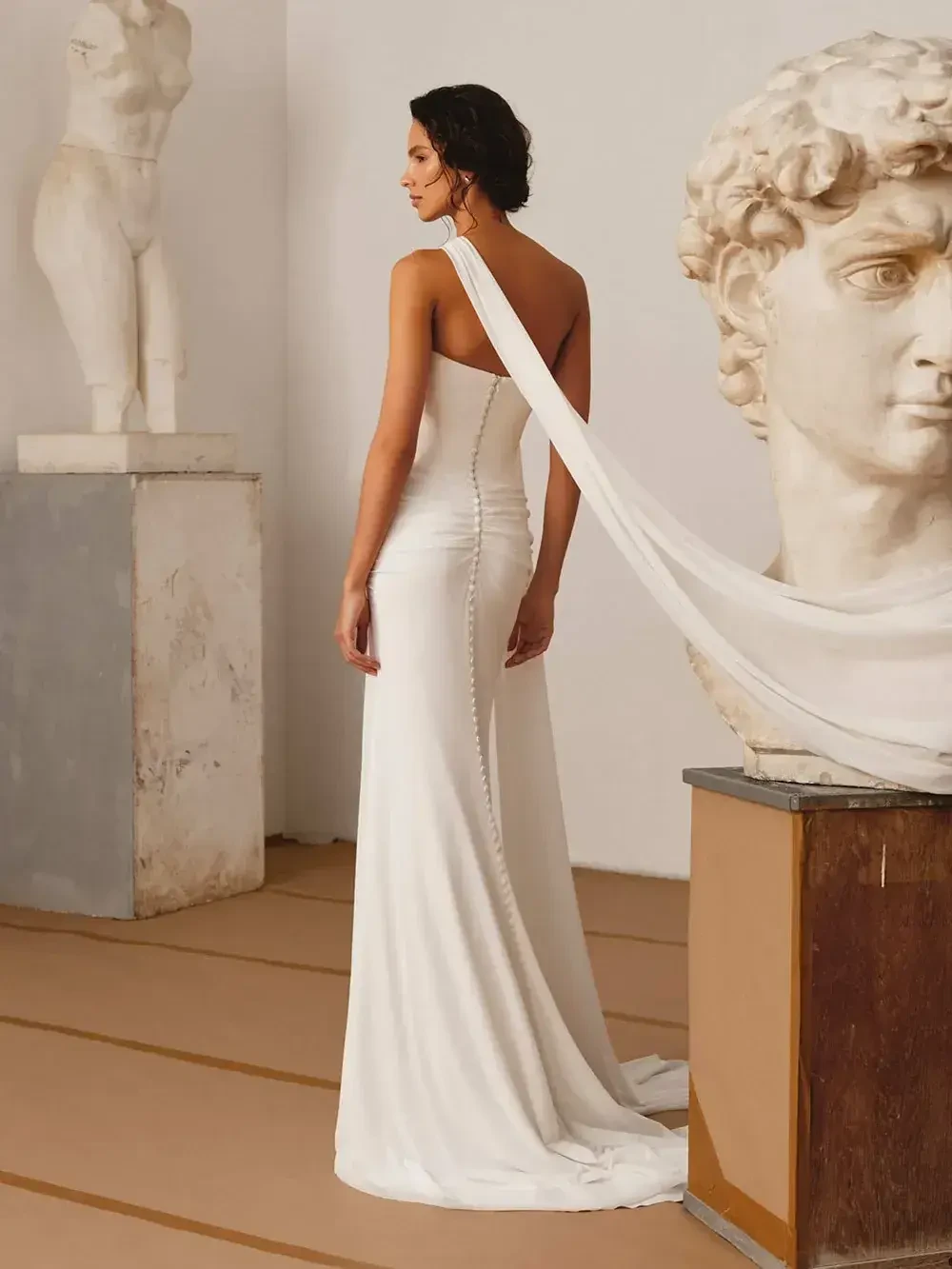 A woman wearing a white gown stands with her back to the camera, draped fabric flowing from her shoulder. In the background, there are classical sculptures, including a bust and a figure.