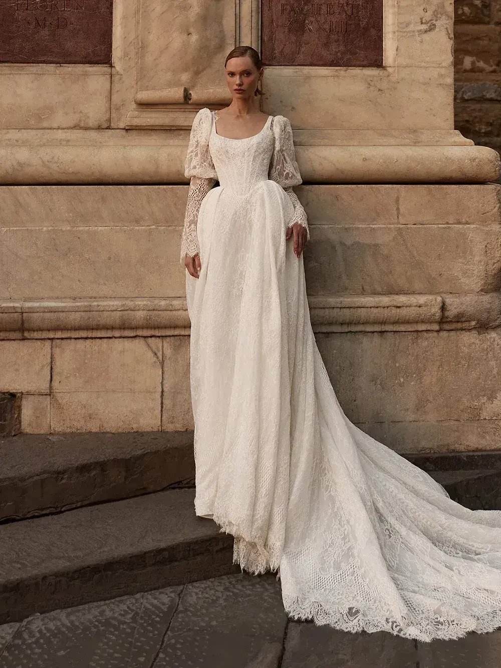 A woman in a flowing white dress with long lace sleeves stands on stone steps against a textured wall.