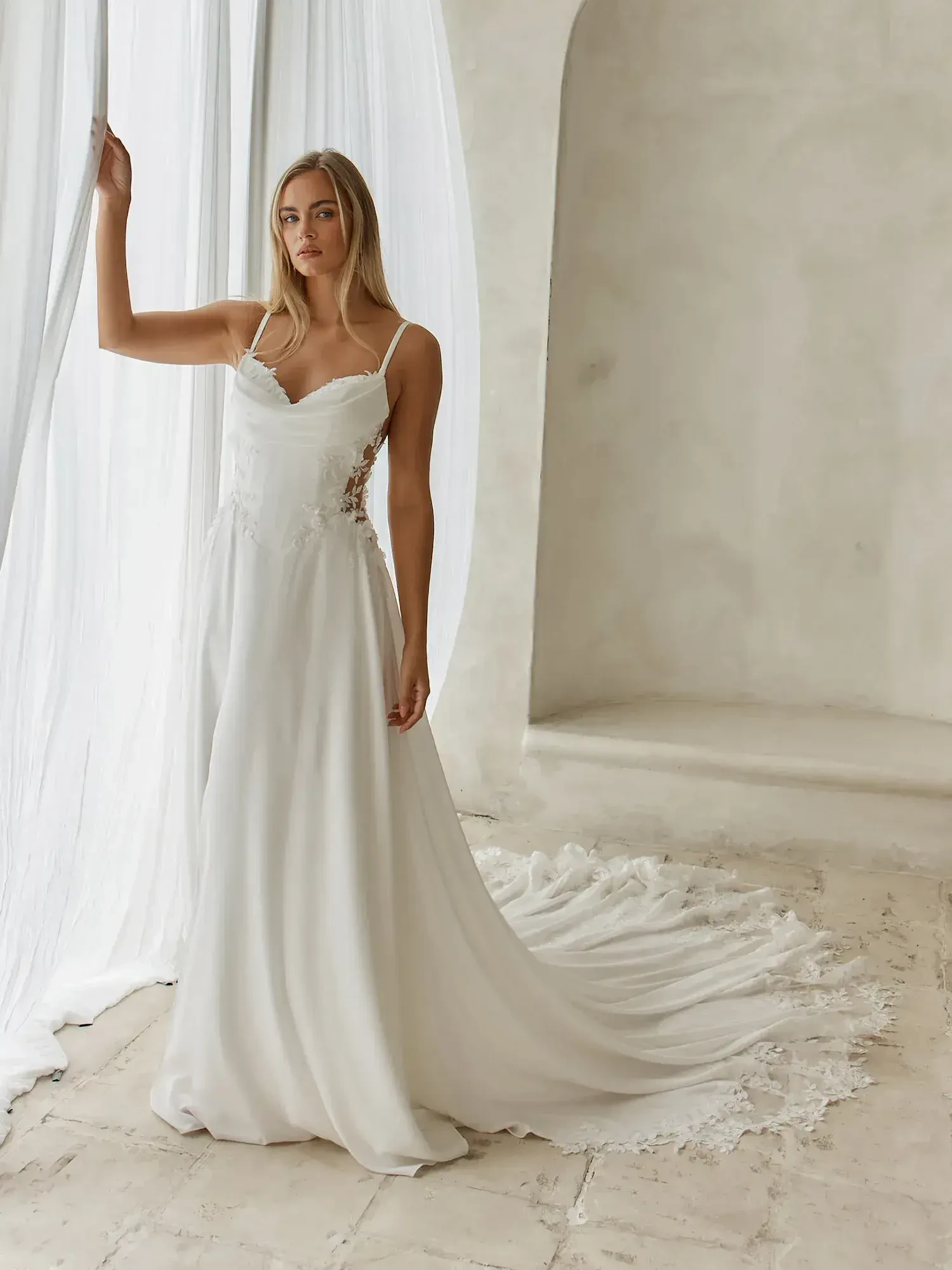 A model is wearing a white wedding dress with thin straps, standing by a sheer curtain in an elegantly styled room.