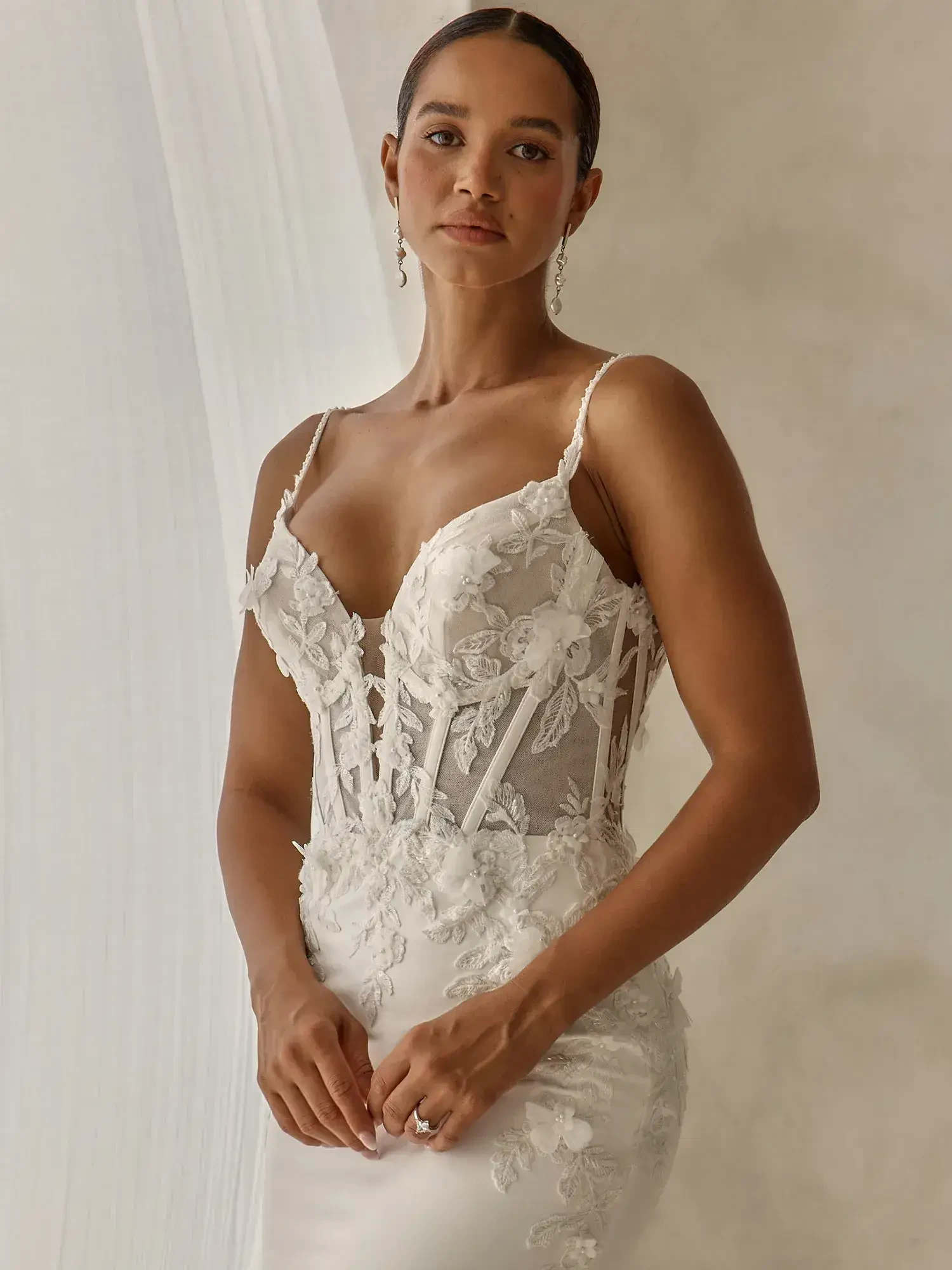A woman in an elegant white bridal gown with intricate lace details and thin straps, standing against a neutral background.
