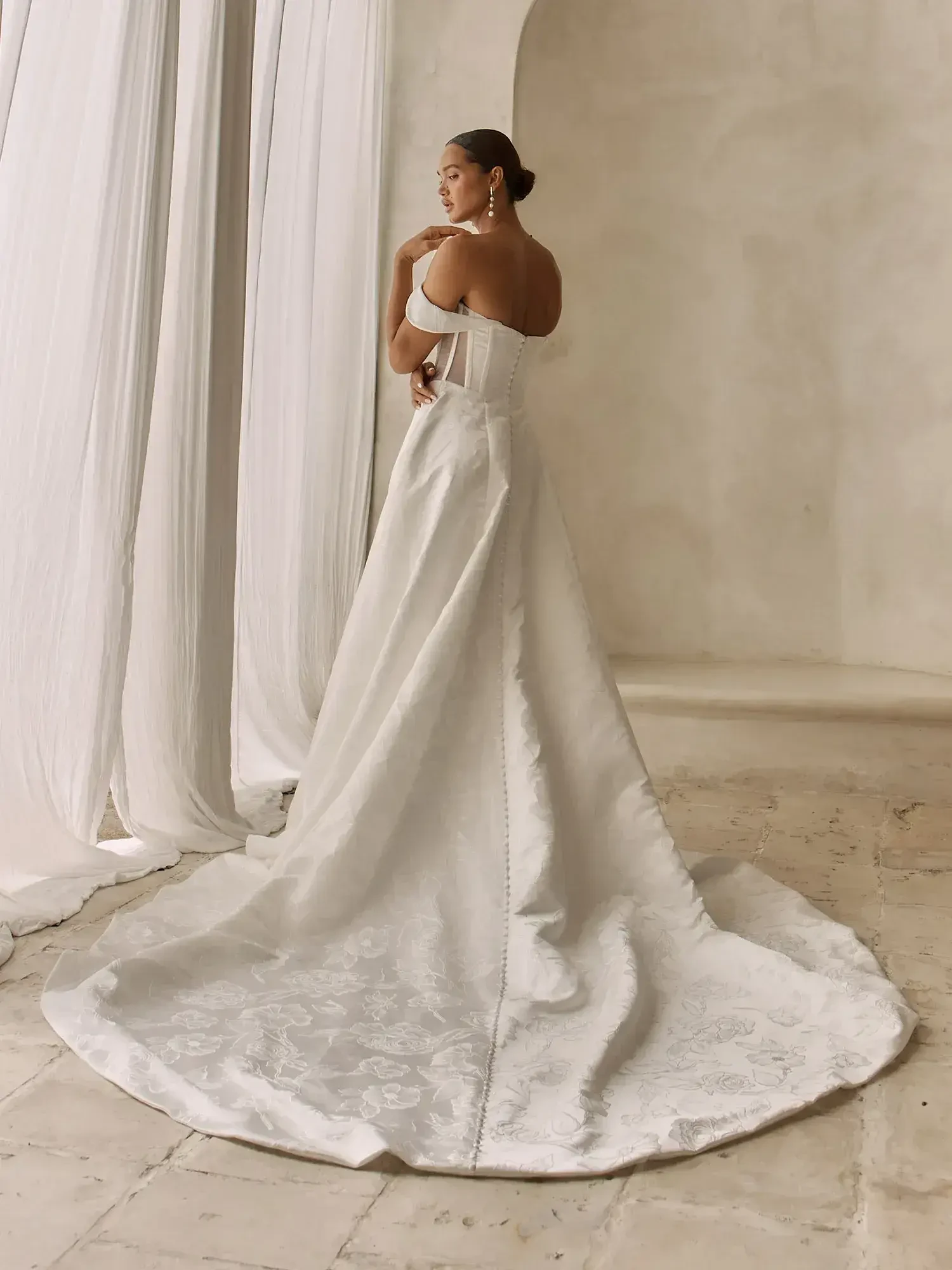 A woman wearing a long, white dress with a train, standing with her back to the camera in a softly lit setting.