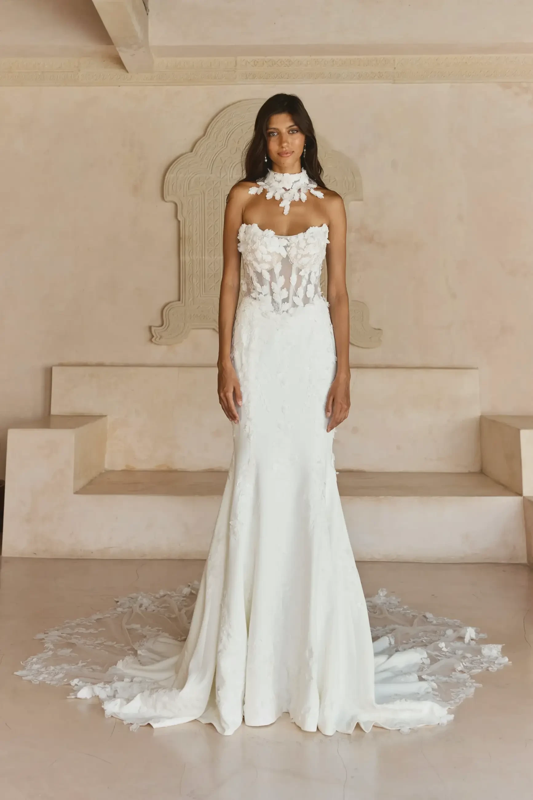 A woman stands in a fitted white wedding gown with intricate floral details at the bodice. The gown has a train and the setting is minimalistic with light-colored walls and floors.