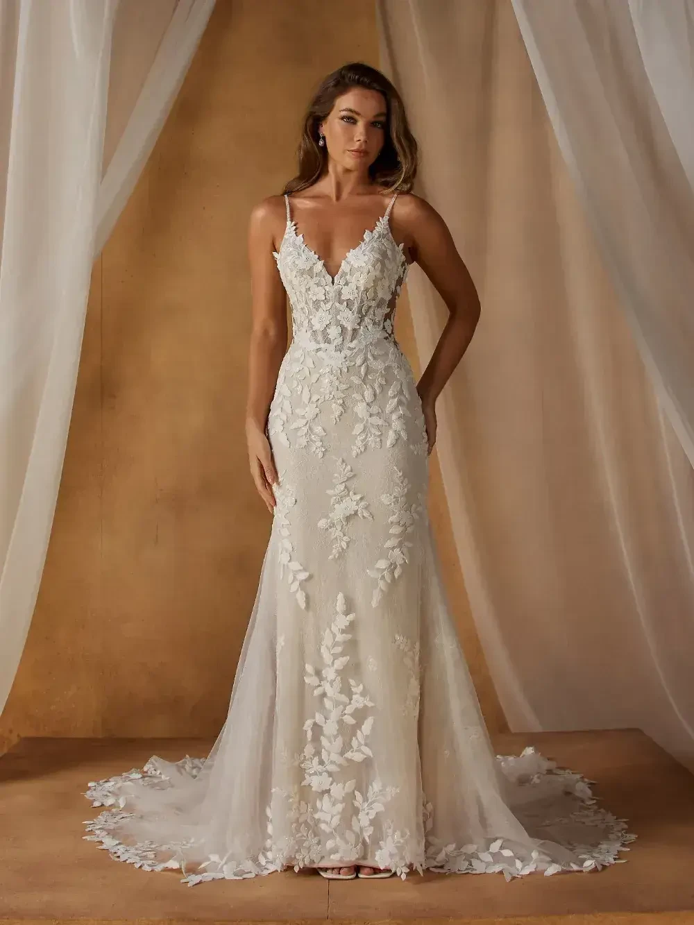 A woman wearing a fitted, elegant wedding dress with lace detailing and a sheer train, standing in front of a soft, draped background.