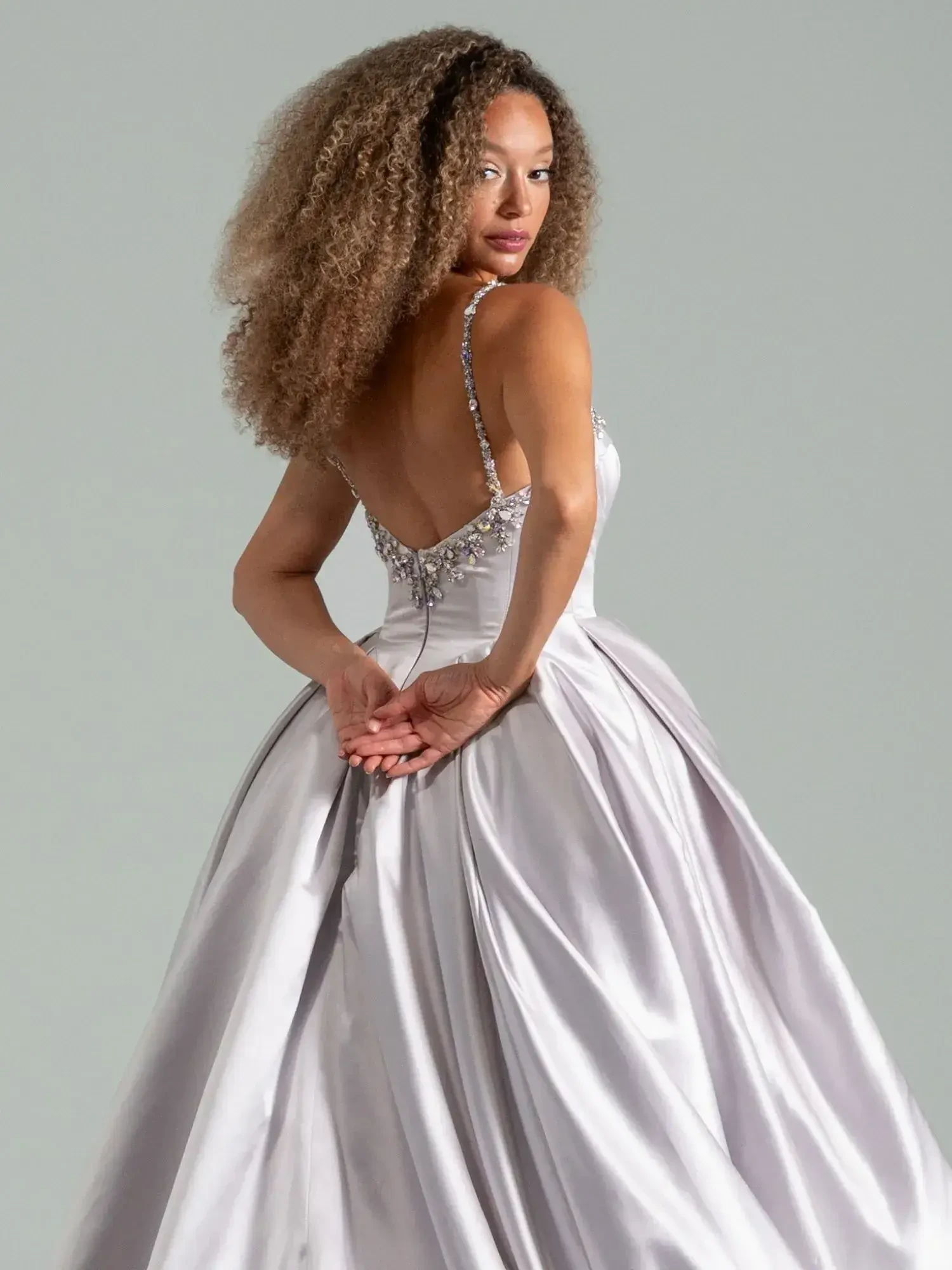 A woman in a silver evening gown turns around, looking over her shoulder with curly hair.