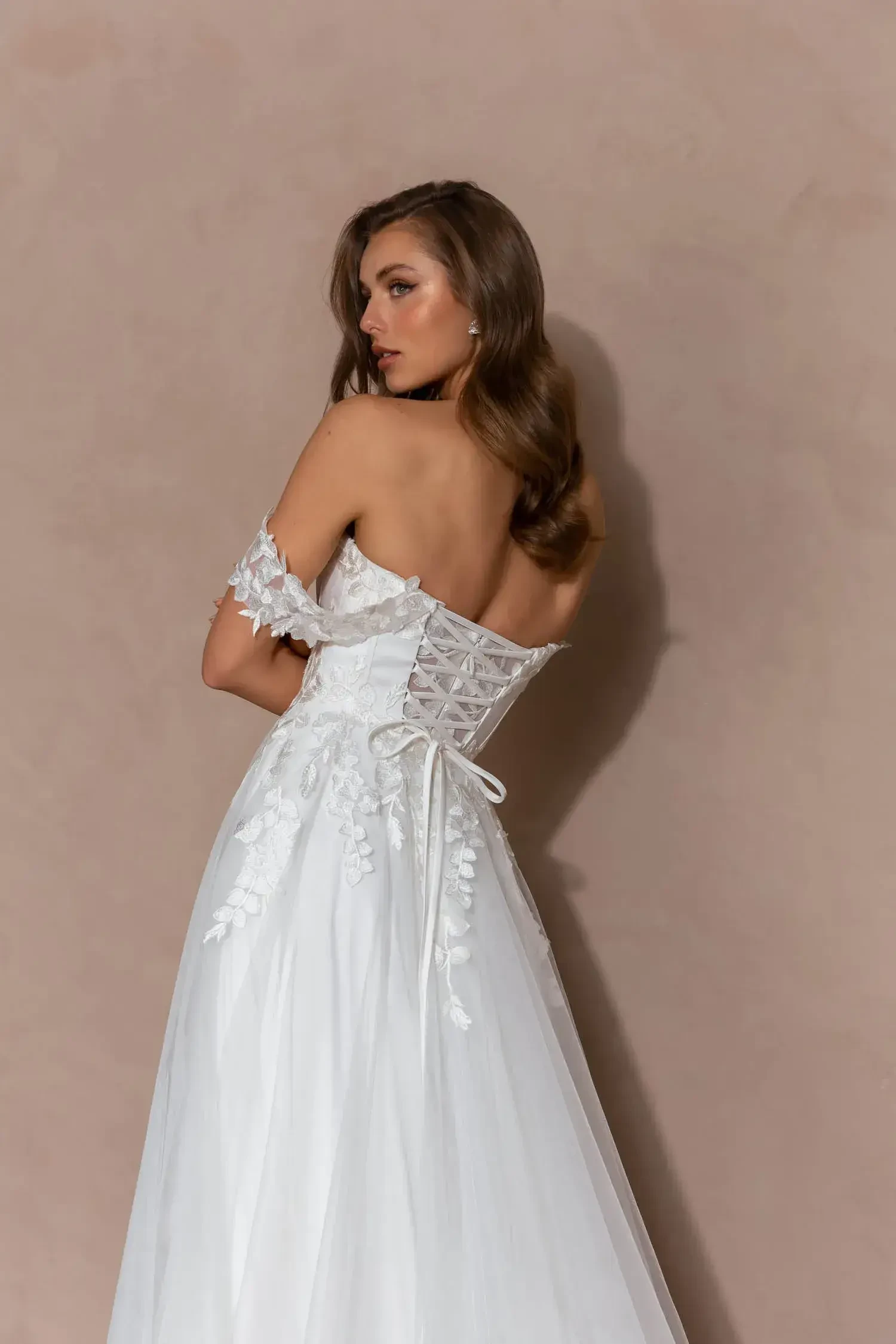 A woman in a white dress is seen from the back, showcasing a lace-up design and floral accents on the gown.