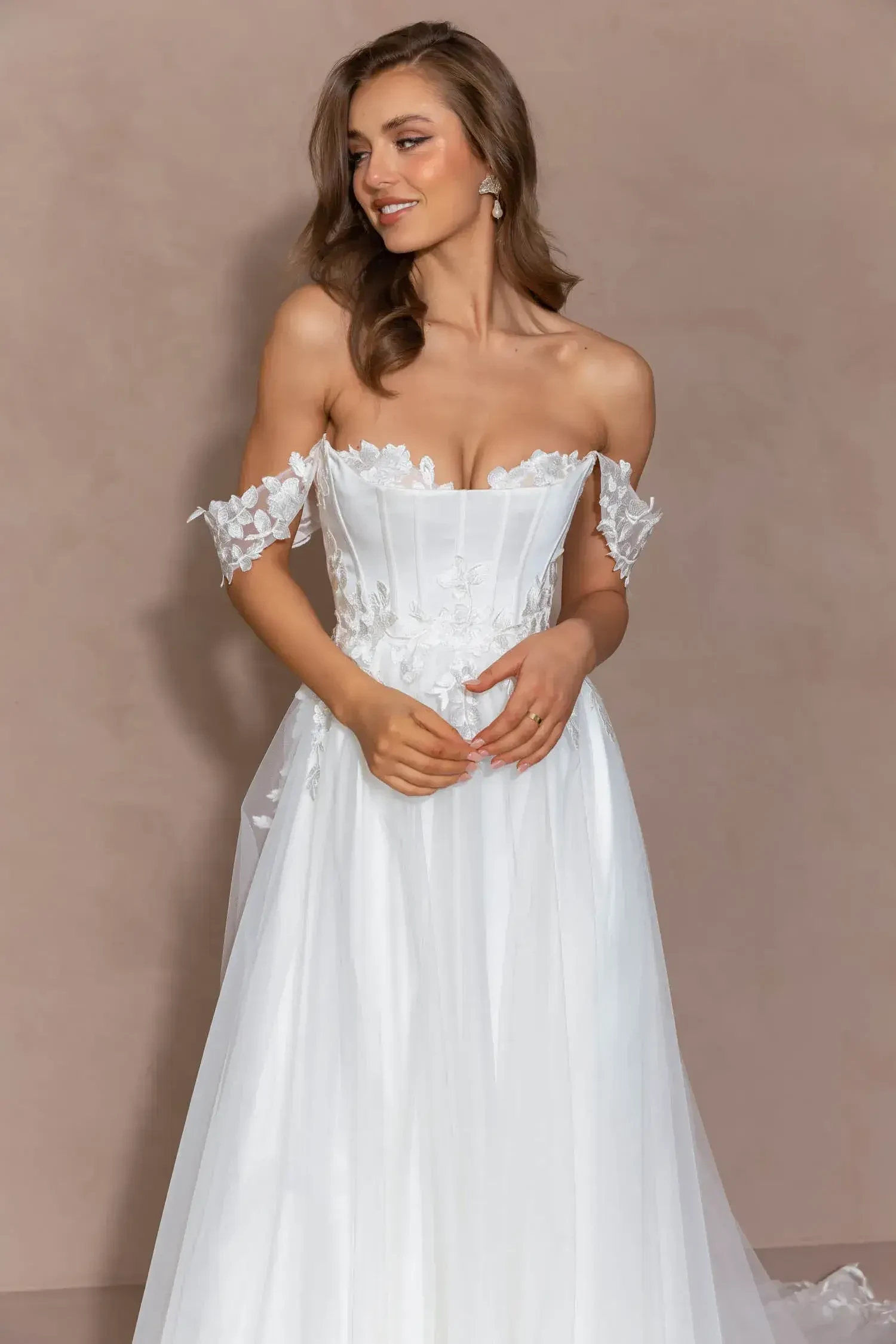 A woman in a white off-the-shoulder wedding dress with floral details, standing against a light brown background. She has long, wavy hair and is smiling softly.