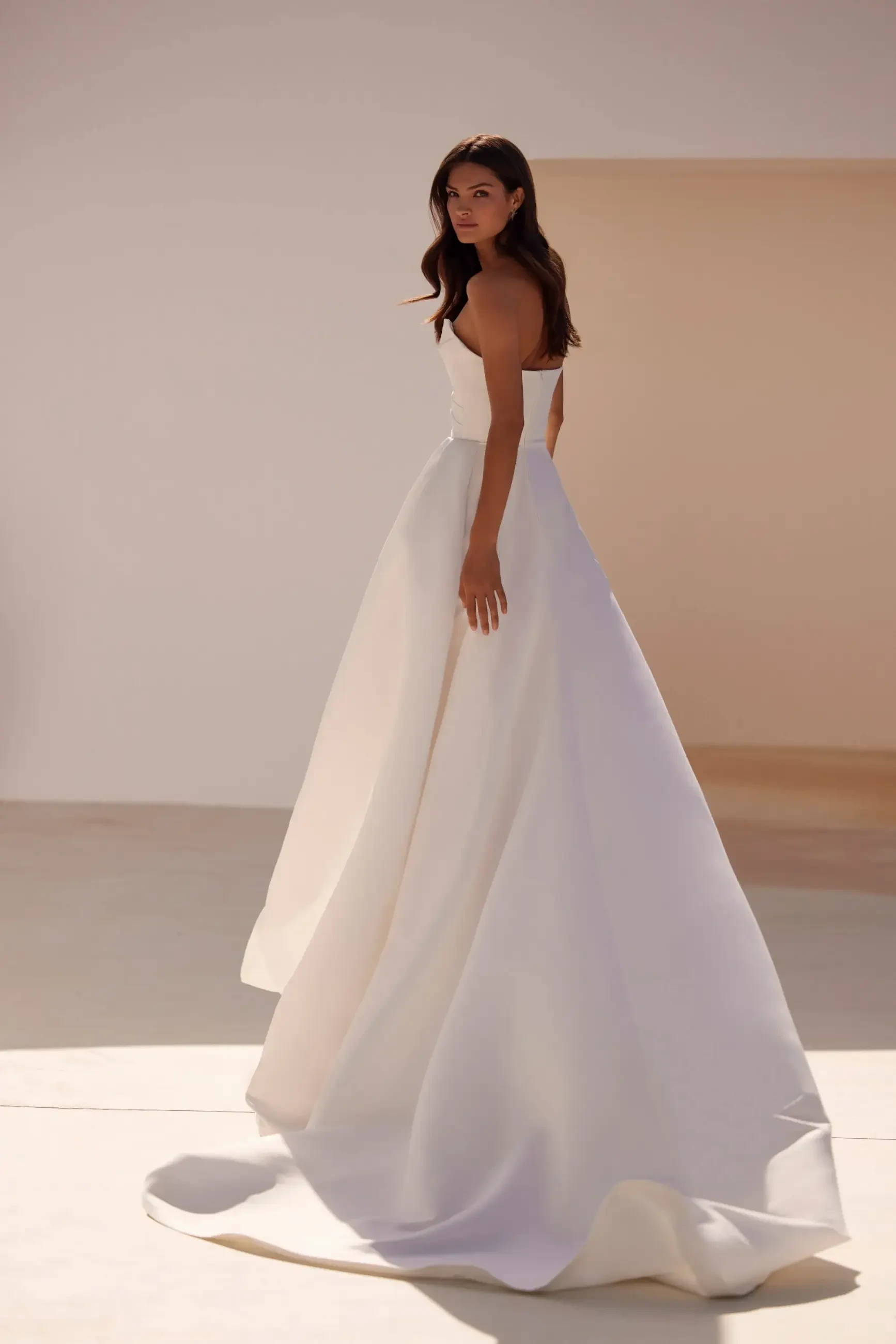 A woman in an elegant, flowing white strapless gown looks over her shoulder in a minimalist, sunlit setting, conveying grace and serenity.