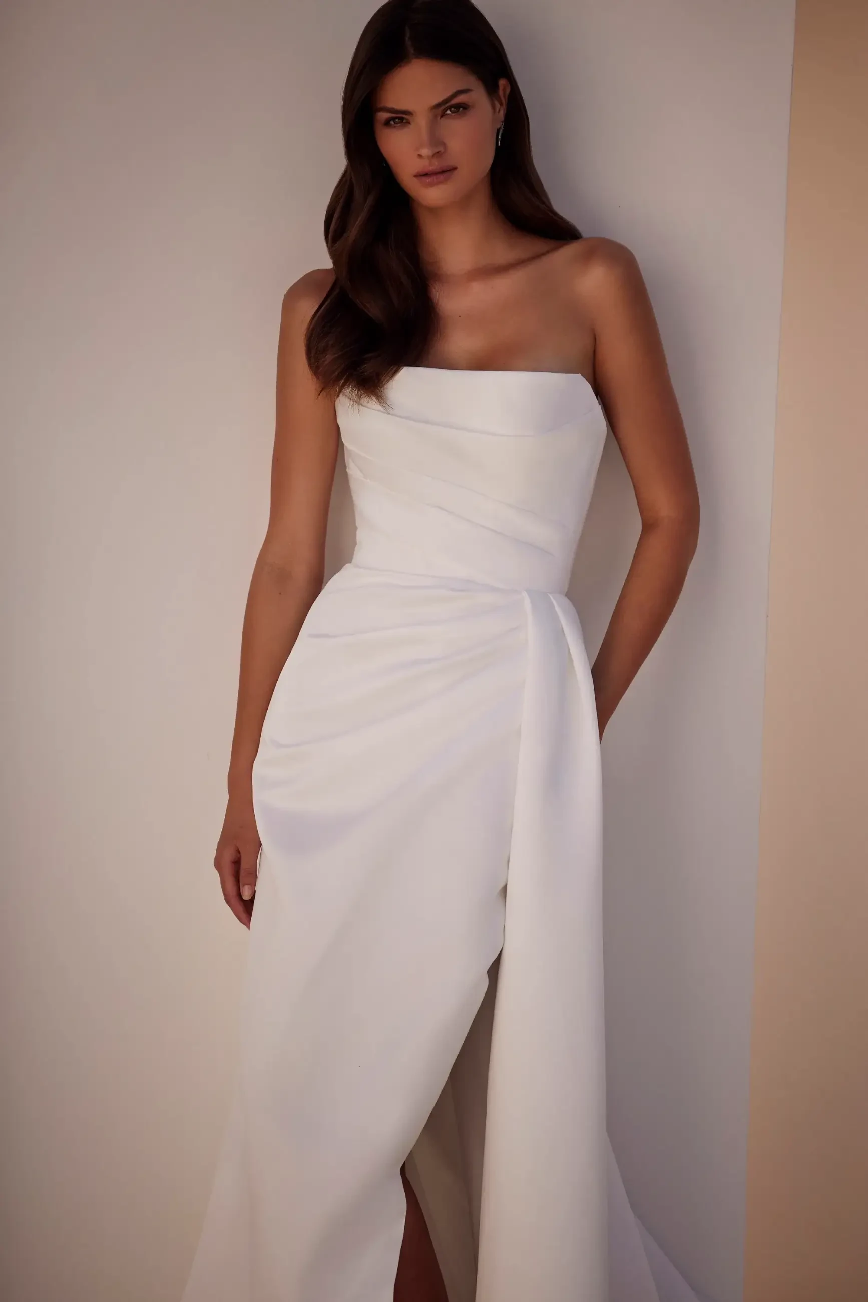 A woman wearing a strapless white dress stands against a plain wall. Her long, dark hair cascades over her shoulder. The tone is elegant and serene.