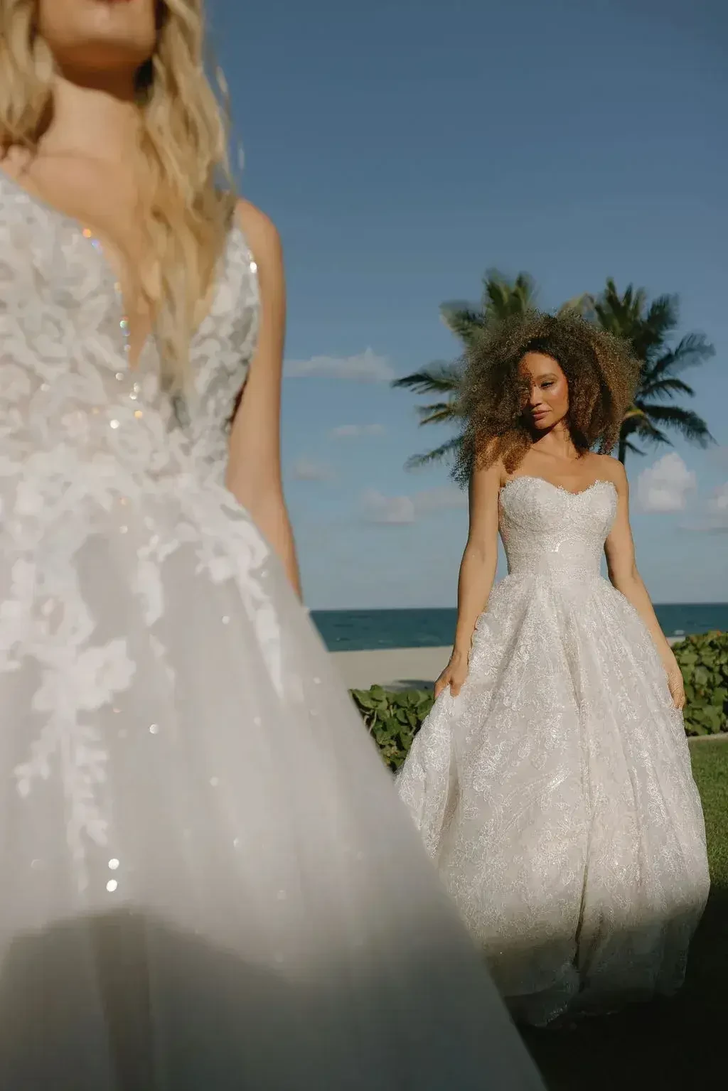 Two women standing outdoors in elegant dresses, with palm trees and ocean in the background. The woman in the foreground is wearing a dress with intricate lace details, while the woman in the background is wearing a shimmering ball gown.
