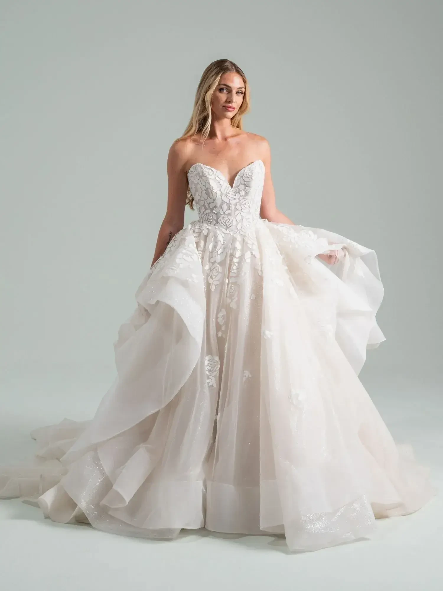 A woman wearing a strapless wedding gown with intricate details and a flowing skirt, posed gracefully against a neutral background.