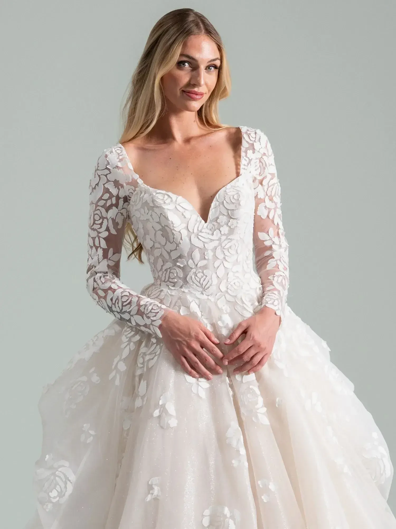 A woman wearing a white wedding dress with long, sheer lace sleeves and floral patterns, smiling at the camera.