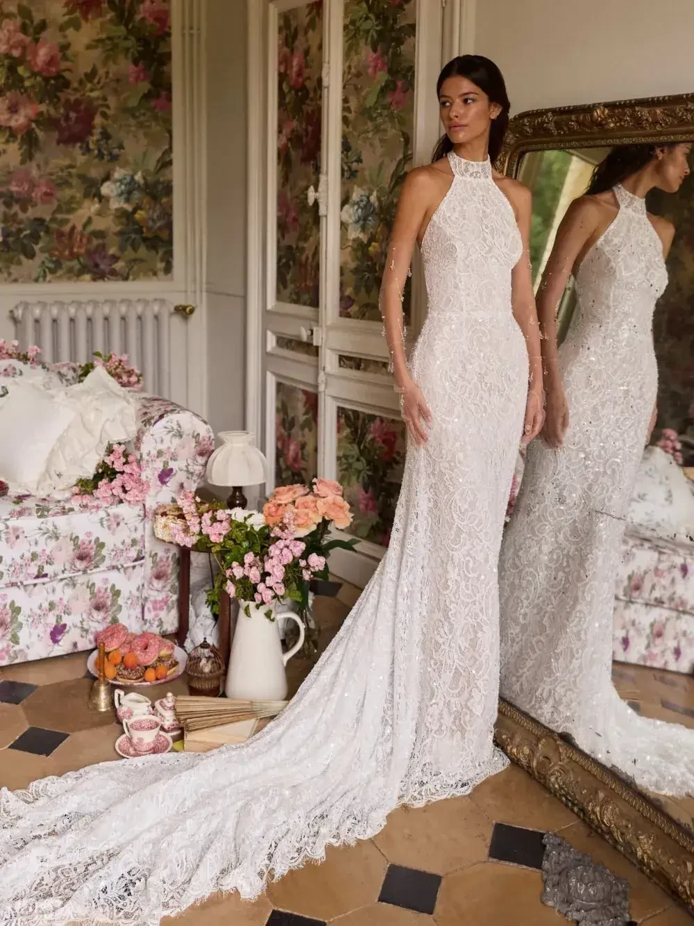 A woman in an elegant white lace wedding dress stands near a large mirror in a floral room. The atmosphere is serene and romantic, with pastel flowers.