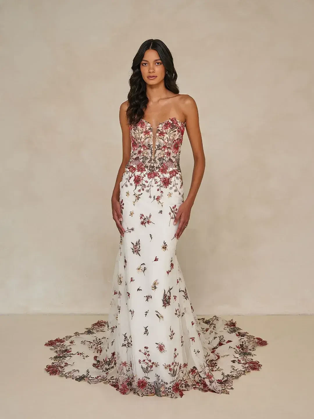 A woman in a strapless white gown adorned with floral embroidery and a long train, standing against a neutral background.