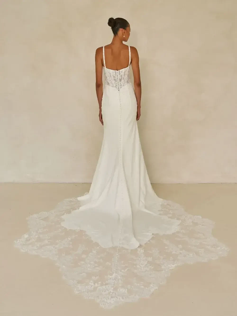 A woman in a white gown stands with her back facing the camera. The dress features spaghetti straps and a long lace train. The setting is neutral.